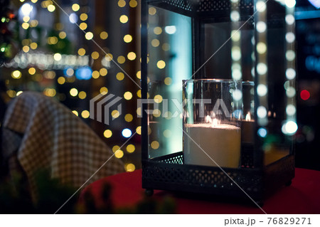 iron lantern with glass inside a candle is burning with a flame of glowing warm light in the night backyard with bokeh from festive garlands on the window in diffocus close-up, nobody. 76829271