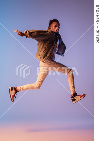 Young stylish man in modern street style outfit isolated on gradient background in neon light. African-american fashionable model in look book, musician performing. 76847058