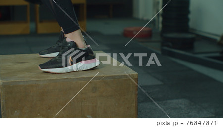 Close up young female with perfect body doing set of box jumps at the gym. Fitness woman in sportswear doing jumps on a big box in a training centre. 76847871