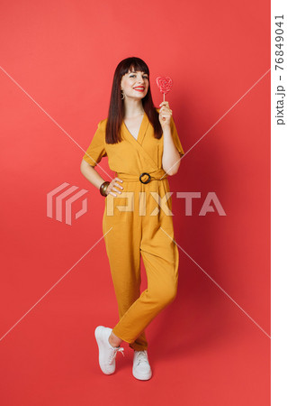 Candy, sweet and emotions concept. Fashion pretty young Caucasian woman in trendy yellow overalls, white sneakers, posing with big red heart shaped lollipop, on bright red studio background Candy, sweet and emotions concept. Fashion pretty young Caucasian woman in trendy yellow overalls, white sneakers, posing with big red heart shaped lollipop, on bright red studio background 76849041