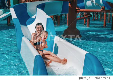 Little girl in swimsuit goes down the blue slides to the pool, Mom and daughter play and swim in the outdoor pool 76877973
