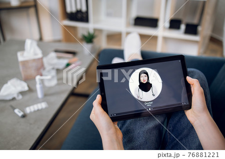 Tablet screen view, close up. Top view of unrecognizable female patient, sitting with digital tablet pc on couch and making video call to young professional Arabian doctor from home Tablet screen view, close up. Top view of unrecognizable female patient, sitting with digital tablet pc on couch and making video call to young professional Arabian doctor from home 76881221
