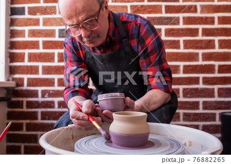 Senior man holding a brush, painting on a ceramic using potter wheel. 76882265