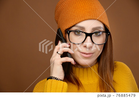 attractive Caucasian white woman wearing glasses in a hat and sweater looking at the camera and talking on the phone. Studio shot on brown background attractive Caucasian white woman wearing glasses in a hat and sweater looking at the camera and talking on the phone. Studio shot on brown background 76885479