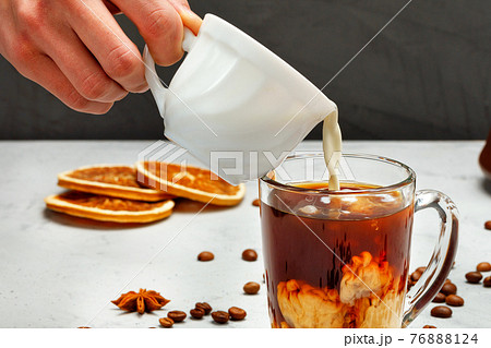 Human hand pours cream into a transparent cup with coffee, close-up. 76888124