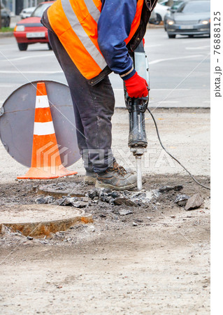 A road worker works with a jackhammer on a cone-fenced section of road. A road worker works with a jackhammer on a cone-fenced section of road. 76888125
