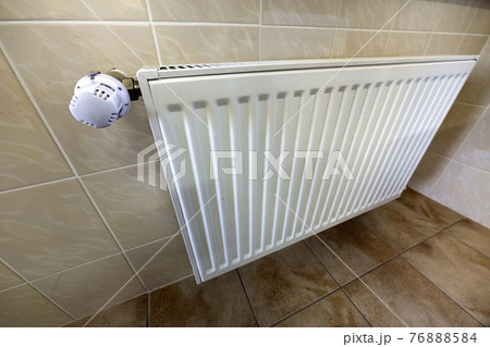 Close-up of white heating radiator with thermostat valve ion tiled walls and floor background. Comfortable warm home interior, climate control, money saving concept. Close-up of white heating radiator with thermostat valve ion tiled walls and floor background. Comfortable warm home interior, climate control, money saving concept. 76888584