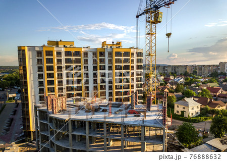 Apartment or office tall building under construction. Brick walls, glass windows, scaffolding and concrete support pillars. Tower crane on bright blue sky copy space background. 76888632