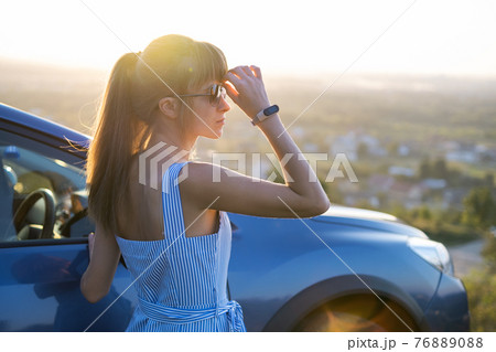 Happy young woman driver in blue dress enjoying warm summer evening standing beside her car. Travelling and vacation concept. Happy young woman driver in blue dress enjoying warm summer evening standing beside her car. Travelling and vacation concept. 76889088