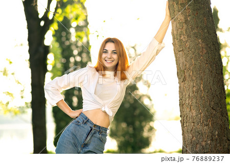Young positive redhead woman in white blouse and blue jeans standing in summer park enjoying warm day. Young positive redhead woman in white blouse and blue jeans standing in summer park enjoying warm day. 76889237