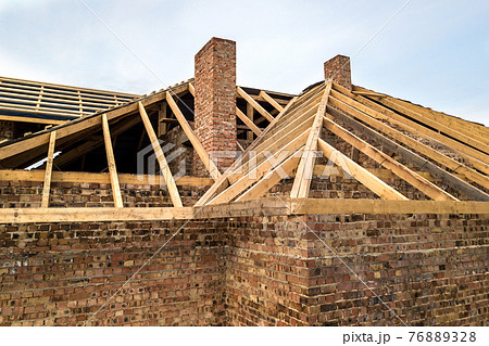 Private residential house with wooden roof frame structure under construction. Unfinished brick building under development. Private residential house with wooden roof frame structure under construction. Unfinished brick building under development. 76889328