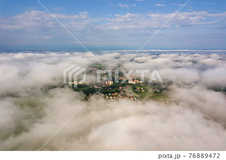 Aerial view of white clouds above a town or village with rows of buildings and curvy streets between green fields in summer. Countryside landscape from above. 76889472