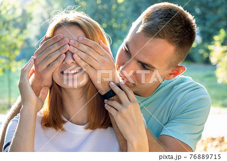 Happy teenage couple having fun together outdoors in sunny summer park surprising one another with closing eyes. Happy teenage couple having fun together outdoors in sunny summer park surprising one another with closing eyes. 76889715