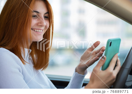 Happy redhead girl texting on her mobile phone behind the wheel driving a car. Happy redhead girl texting on her mobile phone behind the wheel driving a car. 76889771