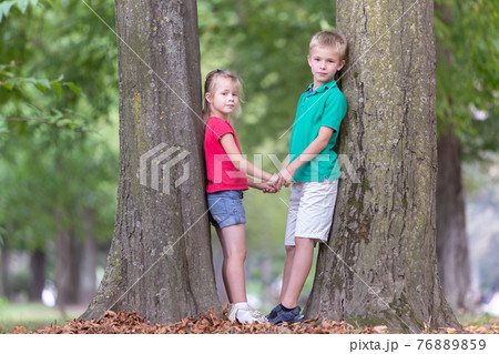 Portrait of two pretty cute children boy and girl standing near big tree trunk in summer park outdoors. 76889859