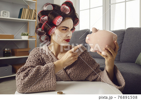 Funny woman in hair curlers shaking finger at piggy bank determined to save up money Funny woman in hair curlers shaking finger at piggy bank determined to save up money 76891508