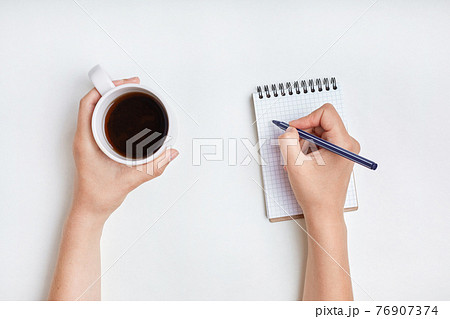 Top view of female hand writes in spiral notebook, takes notes with cup of tea. Woman writes in her diary. Beautiful female's hand holds pen above notebook paper with mug of coffee 76907374