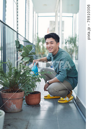 Attractive Young Man on Apartment Balcony Watering Plants in Box from Blue Watering Can Attractive Young Man on Apartment Balcony Watering Plants in Box from Blue Watering Can 76910306