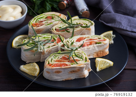 Rolls of thin pita bread and red salted salmon with lettuce leaves on a black ceramic plate, dark wooden background. 76919132