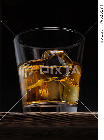 Glass of single malt whiskey with ice cubaes on top of wooden board on black. 76920184
