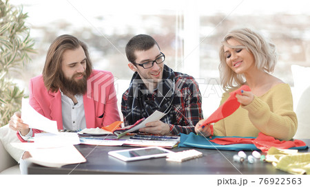 Smiling tailor businesswoman sitting at sewing machine and making handmade clothing while marketing assistant working on new projec 76922563