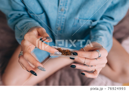 Woman wearing jeans shirt making hand rolled cigarette with tobacco at home 76924993