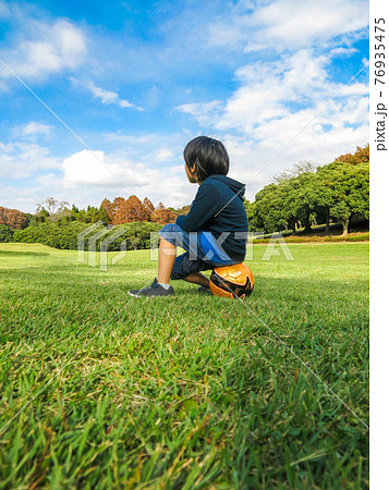 晴れた日に芝生の公園でボールに座るサッカー少年 76935475