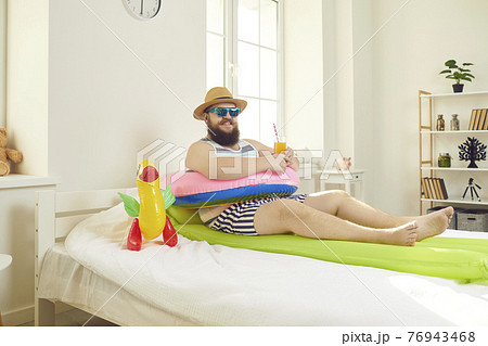 Funny bearded man with travel accessories lying in bed imagining resort vacation Funny bearded man with travel accessories lying in bed imagining resort vacation 76943468