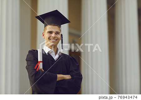 Successful graduate in academic gown posing with diploma in hands against university background. Successful graduate in academic gown posing with diploma in hands against university background. 76943474