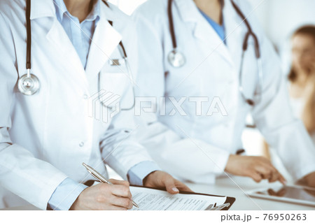 Unknown woman-doctors at work with patient at the background. Female physicians filling up medical documents or prescription while standing in hospital reception desk, close-up. Health care concept Unknown woman-doctors at work with patient at the background. Female physicians filling up medical documents or prescription while standing in hospital reception desk, close-up. Health care concept 76950263