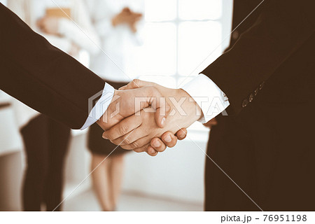 Businessman and woman shaking hands with colleagues at the background. Handshake at meeting in office. Concept of success in business Businessman and woman shaking hands with colleagues at the background. Handshake at meeting in office. Concept of success in business 76951198
