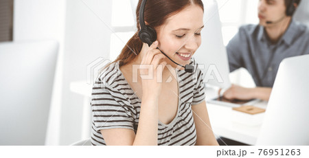 Casual dressed young woman using headset and computer while talking with customers online. Group of operators at work. Call center, business concept 76951263