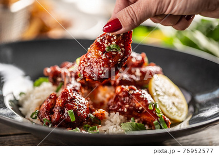 Woman's hand holding a sticky glazed spicy chicken with sesame seeds with a plate with rice in the background 76952577