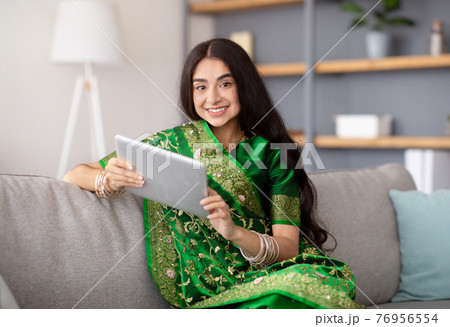 Cheerful Indian lady in traditional saree sitting on cozy sofa with tablet pc attending online meeting from home Cheerful Indian lady in traditional saree sitting on cozy sofa with tablet pc attending online meeting from home 76956554