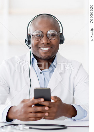 Hot line medical center. Friendly black male doctor in headphones holding smartphone in hand 76956620