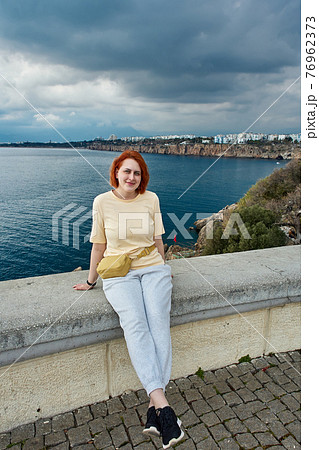 Young smiling redhead Caucasian woman posing for photographer while sitting on fence of observation point overlooking sea in Antalya. Young smiling redhead Caucasian woman posing for photographer while sitting on fence of observation point overlooking sea in Antalya. 76962373