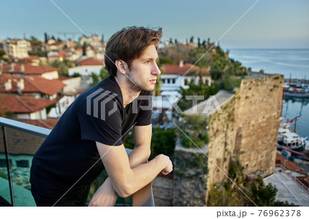 Slender European man, about 20 years old, sadly looks at old city in Antalya from observation point. Slender European man, about 20 years old, sadly looks at old city in Antalya from observation point. 76962378