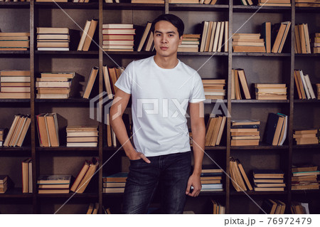 Asian man in white T-shirt stands against the background of bookshelves. Mockup. 76972479