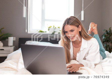 Smiling girl lays on the bed in bedroom and use laptop for remote work Smiling girl lays on the bed in bedroom and use laptop for remote work 76974349