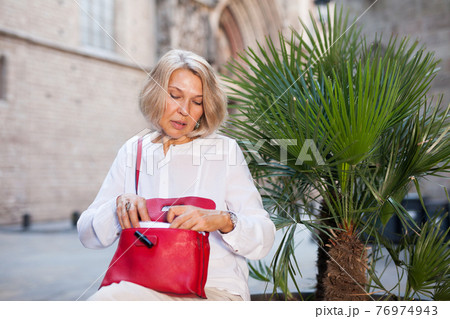 Elderly woman is looking for something in her handbag 76974943