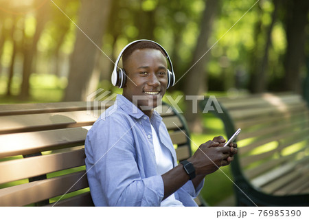 Handsome black man relaxing on bench in park and enjoying his favorite music on smartphone 76985390