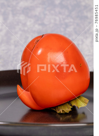 One ugly orange persimmon on black plate on light grey backdrop close up. Zero waste food. Reasonable sustainable consumption. Persimmon - source of vitamins and trace elements. Vertical orientation. 76985451