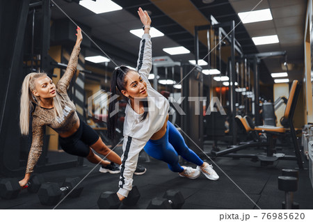 Two beautiful women with athletic figures perform a side plank pose in gym. Two beautiful women with athletic figures perform a side plank pose in gym. 76985620