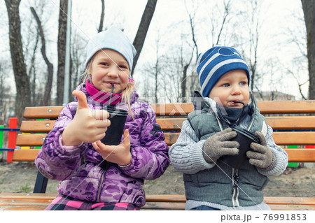 Two cute adorable siblings children sitting on wooden bench and drinking hot chocolate, tea or cocoa from paper cups during walk at city street park or backyard outdoors. Brother and sister enjoy fun Two cute adorable siblings children sitting on wooden bench and drinking hot chocolate, tea or cocoa from paper cups during walk at city street park or backyard outdoors. Brother and sister enjoy fun 76991853