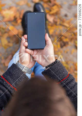 Man holding smartphone outdoors with yellow autumn leaves on the ground Man holding smartphone outdoors with yellow autumn leaves on the ground 77008226