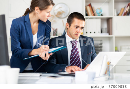 Female assistant writing down tasks on clipboard near boss 77009858