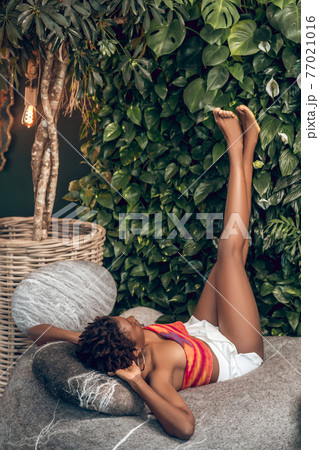 Dark-skinned young woman in a bright top and white shorts laying with her legs up Dark-skinned young woman in a bright top and white shorts laying with her legs up 77021016