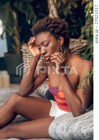 Dark-skinned young woman in a bright top and white shorts looking thoughtful Dark-skinned young woman in a bright top and white shorts looking thoughtful 77021017