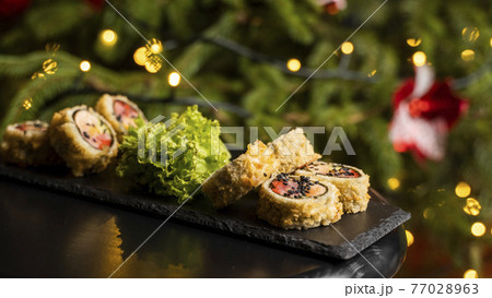 Custom sushi roll in tempura with nori, fresh salmon, tuna, avocado, masago caviar, drizzled with pineapple sauce with salad pouring as decoration on a black plate on a wooden table for Christmas. 77028963