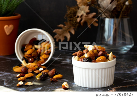 Mix of delicious assorted nuts and dried fruit in white cups on a table, sunny morning. Healthy gourmet vegetarian natural snack. 77030427